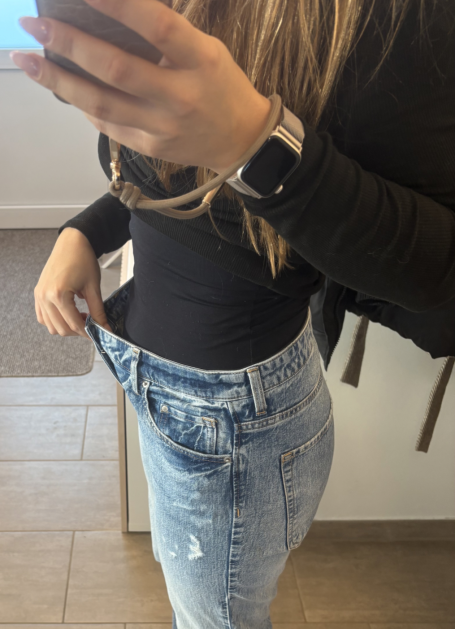 Junge Frau in enger Bluse und Jeans hält die Hose am Bund und schaut in einen Spiegel.